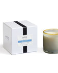 A Lafco NY Beach House Candle made of soy wax sits in a hand-blown glass vessel with a gold rim, next to its white box featuring a black stripe and blue scent label.