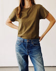 A person stands against a plain background wearing Hey Gang's The Crew, a brown 100% cotton vintage crew t-shirt, paired with blue jeans. One arm rests on their hip, and their head is slightly out of frame.