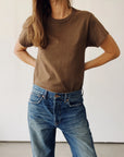 Wearing "The Crew" vintage brown t-shirt by Hey Gang and blue jeans, a person stands hands on hips against a plain light background. Their face is partly out of frame, with hair draped over one shoulder.