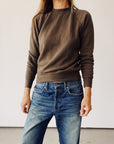 A person wears The Sweatshirt by Hey Gang in brown with blue jeans, standing against a plain light background; their face is partially out of frame.