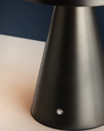 A close-up of the Hudson Valley Hopkins Rechargeable lamp base, featuring a modern metallic cone shape with a small round button, set against a contrasting dark and light background.
