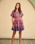 A woman poses on a wooden floor in the Hunter Bell NYC Howard Dress—a colorful, short-sleeved, knee-length Tapestry Floral dress with an elastic waist and brown flats—in front of a beige circular backdrop.