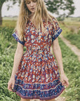 A woman with long blonde hair wears the Howard Dress by Hunter Bell NYC, featuring a shirred skirt, as she stands on a dirt path surrounded by green grass and trees on a cloudy day.