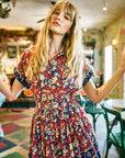 A woman with long blonde hair wears the Howard Dress by Hunter Bell NYC—a red tapestry floral-patterned dress with an elastic waist—standing indoors in a vibrant vintage-inspired room with green chairs and patterned floor tiles.