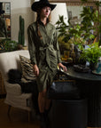 A woman wears The Wrap Front Dress by THE SHIRT in olive green, styled with a black hat. She stands indoors beside plants and glassware near a beige armchair and a large black bag on a round stool.