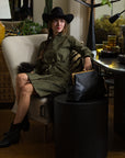 A woman wears THE SHIRT’s The Wrap Front Dress—an olive-green, long-sleeve wrap dress—styled with a black hat and ankle boots, as she sits on a cream armchair. A black handbag rests on the nearby round table in the eclectic room.