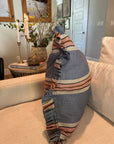 The Rennah Collective Foxy Blue Striped Ruffle Pillow 22x22 sits on a beige sofa in a cozy living room, surrounded by candles, books, flowers, and framed pictures for a welcoming touch.