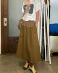 Indoors, a person models the Aquarius Cocktail Le Cargo Skirt with large pockets, paired with a graphic tee featuring sheer pink at the neckline, cream and brown cowboy boots, and a woven bag. Rolls of paper appear in the background.