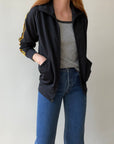 A person wears the Red Vintage Co. 1980s Black & Yellow Track Jacket with yellow stripes and raglan sleeves, paired with a gray top and blue jeans, standing against a plain light background with hands in the jacket pockets.