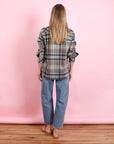 A person with long blonde hair stands facing away, wearing the BCS Group Lumber L/S Shirt, blue jeans, and brown sandals, against a pink background with a wooden floor.
