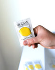 A hand holds a Herban Essentials Essential Oil Towelettes packet featuring lemon imagery, emphasizing its refreshing lemon scent; two more towelette packets are blurred in the background on a white surface.