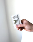 A hand holds a packet of Herban Essentials Essential Oil Towelettes, featuring a white design with the product name and an illustration of a eucalyptus branch on the front.