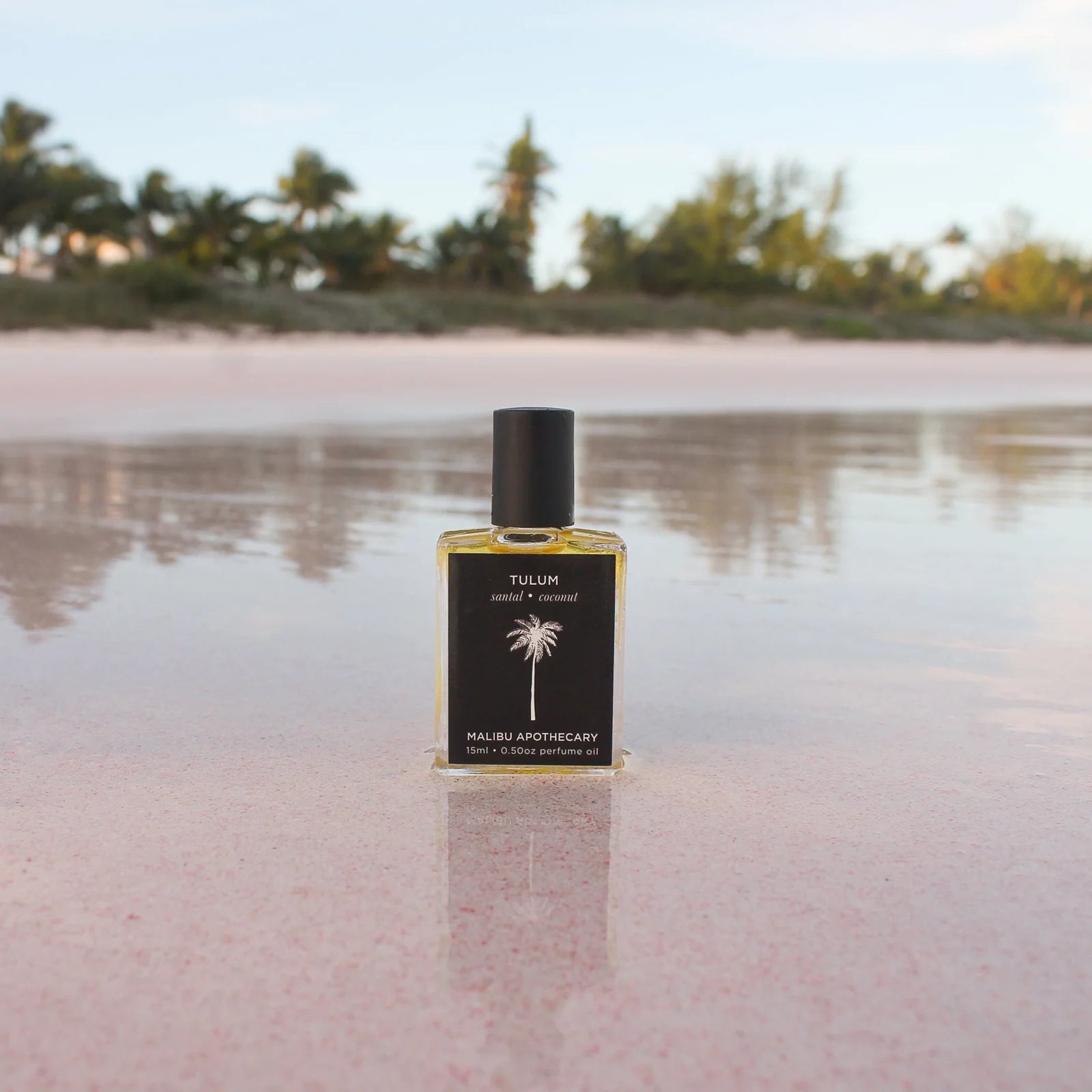 A bottle of "Soleil Roller Parfum" by Faire is placed on a reflective wet sand at sunset, with pink hues in the background and a palm tree silhouette on the bungalow-style bottle.