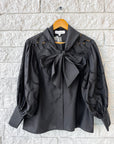 The Shay Blouse by Hunter Bell NYC, featuring black eyelet fabric, roomy sleeves, a button-down front, and a standout bow collar, is displayed on a wooden hanger against a white brick wall.