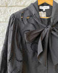 Close-up of the Shay Blouse by Hunter Bell NYC—a black eyelet top with spacious sleeves and a dramatic bow collar, hanging on a wooden hanger against a textured light stone wall.
