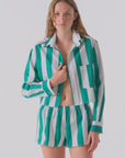 A woman models Cissa's The Boxer set—green and white vertical stripes in soft Egyptian cotton—with a partially unbuttoned shirt, standing against a plain backdrop.