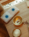 An Indaba 1-1234 Forged Brass Bowl showcases jewelry, like a sun-shaped ring, on a wooden table beside a white crocheted pouch with blue flowers, near a sunny window—stylish and elegant for desk organization.