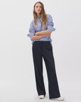 A woman with long hair stands against a white background, arms crossed, wearing Rag & Bone's irina ponte tailored pant in dark fabric and a blue-and-white striped button-up shirt, looking at the camera.