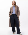 A woman wears Rag & Bone’s irina ponte tailored pant in navy, paired with white shoes, a blue-and-white striped button-up shirt, and a long taupe coat, standing against a plain white background.