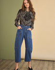 A woman stands with hands in pockets, wearing wide-leg blue jeans, black shoes, and the Frances Blouse by Hunter Bell NYC—a versatile black blouse with a neck bow—against a beige and green backdrop.