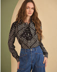 A woman with long wavy brown hair wears the Hunter Bell NYC Frances Blouse, a black patterned long-sleeve top, and high-waisted blue jeans, standing in front of a beige and green background.