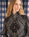 A woman with long blonde hair wears the Frances Blouse by Hunter Bell NYC—a black blouse with a white paisley bandana pattern—standing before blue and white checkered curtains, gazing calmly to the side.