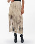 A person wears the Rag & Bone jasmine fringe skirt—a beige, high-waisted suede skirt with long fringe—paired with black knee-high boots, standing against a plain white background.