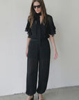A woman in sunglasses and black pleated wide-leg pants wears the Le Superbe Josephine Blouse—sheer, lightweight silk with a neck tie and ruffled sleeves—with black heeled sandals, standing against a plain white wall.