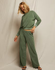A person with shoulder-length blonde hair wears the Perfectwhitetee Saylor Loop Terry Sweatshirt in sage green with matching wide-leg pants and black sandals, standing against a plain fabric backdrop.