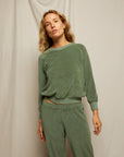 A woman with wavy blonde hair stands against a neutral fabric backdrop, wearing the Perfectwhitetee Saylor Loop Terry Sweatshirt in matching sage green with long sleeves and soft loop terry fabric pants.