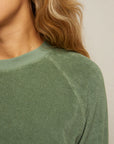 Close-up of a person with long, wavy blonde hair wearing the Perfectwhitetee Saylor Loop Terry Sweatshirt in sage green, showing only the side of the face, neck, shoulder, and upper chest against a plain background.