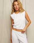A woman with wavy blonde hair wears the ELLIE terry banded tank by Perfectwhitetee and matching white pants, standing before a neutral, textured backdrop.