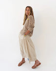 A woman with long brown hair stands sideways against a white background, wearing the Kerri Rosenthal Summerland V Neck Sweater, a beige ankle-length skirt, brown sandals, and a yellow watch. Her hands are in her skirt pockets.