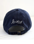 The Kerri Rosenthal Baseball Hat Heart Patch features "loved" and a white embroidered heart above the adjustable strap—a navy cap and chic essential, shown here on a light surface.