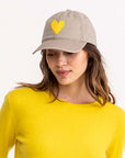 A woman smiles softly, looking down while wearing a Kerri Rosenthal Baseball Hat Heart Patch and a bright yellow sweater against a plain white background.