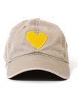 The Kerri Rosenthal Embroidered Heart Hat features a beige design with a yellow heart stitched on the front, offering a fresh take on classic collegiate style. Shown against a plain white background.