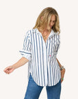 A woman models the Kerri Rosenthal Mia Mr Sharpie Stripe Shirt—white with navy blue stripes—paired with blue jeans and layered necklaces, standing with one arm slightly raised against a neutral background.