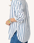 A woman wears the Kerri Rosenthal Mia Mr Sharpie Stripe Shirt—white with vertical blue stripes and rolled sleeves—paired with blue jeans, chunky bracelets, and a ring. Only her upper body is visible from the side.