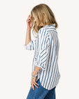 A woman stands sideways, touching her wavy blonde hair while wearing the Kerri Rosenthal Mia Mr Sharpie Stripe Shirt—a women’s cotton poplin shirt with vertical blue stripes and rolled-up sleeves—paired with blue jeans against a plain white background.