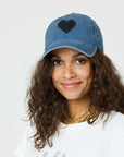 A woman with long curly hair wears the Kerri Rosenthal KR Imperfect Heart Hat, 100% cotton in blue with a black heart patch, paired with a white shirt and gold chain necklace, against a plain light background.