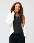 A woman with curly hair smiles while wearing the KR Tank by Kerri Rosenthal, a black cotton tank with an embroidered heart, paired with black pants and a white cardigan draped over her shoulders against a plain background.