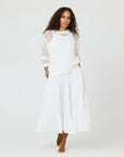 A woman with curly hair stands barefoot against a plain background, wearing the Kerri Rosenthal Catch Open Stitch Sweater—a white, 100% cotton crew neck—paired with a flowing white skirt.