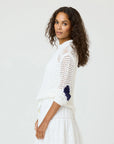 A woman with long curly hair models the Kerri Rosenthal Catch Open Stitch Sweater, featuring blue sleeve detailing, paired with a white skirt. She stands sideways against a plain light background, looking at the camera.