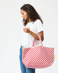 A woman with curly hair in a white t-shirt and blue jeans holds the Kerri Rosenthal KR Heart in Check Tote, a large cotton canvas bag with a bold red-and-white pattern, against a plain white background.
