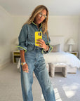 A woman with long hair and glasses takes a mirror selfie in a bright, modern bedroom, wearing the Kerri Rosenthal Pia Denim Western Shirt and high-waisted jeans.