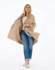 A woman with long blonde hair stands against a white background, wearing the Kerri Rosenthal Cali Fringe Cardigan Coat over a white top, blue cuffed jeans, and white heels. She is looking slightly to the side.