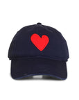 The Embroidered Heart Hat by Kerri Rosenthal is a navy blue baseball cap with a vibrant red heart patch embroidered on the front center.