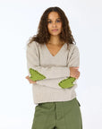 A woman with long curly hair, arms crossed, models the Kerri Rosenthal Frenchy Sweater—beige cashmere with green heart patch elbows—and green pants against a plain white background.