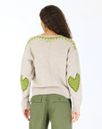 A person with long curly hair is seen from the back wearing the Kerri Rosenthal Frenchy Sweater in beige, featuring heart patch elbows and green whipstitch details, paired with olive green pants against a plain white background.