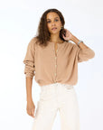 A woman with wavy brown hair wears the beige Kerri Rosenthal Bassett Cardigan and white high-waisted pants, standing against a plain white background with her left hand touching her head.